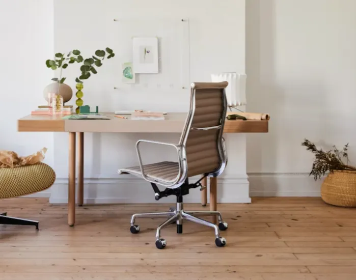 Brown leather Eames chair at wooden desk in bright room with plant